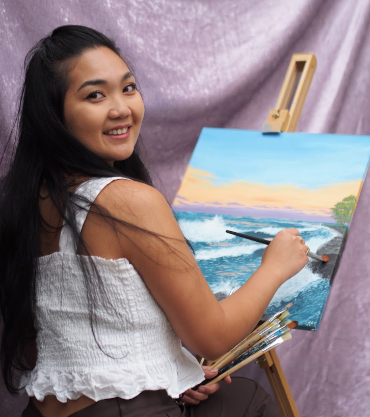 Filipino girl turned back towards a canvas of a seascape painting and a paintbrush on the piece with her head looking and smiling at the camera